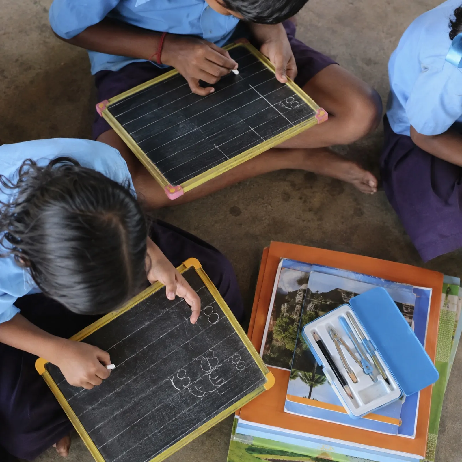 Children writing on chalk slates beside notebooks and a geometry box